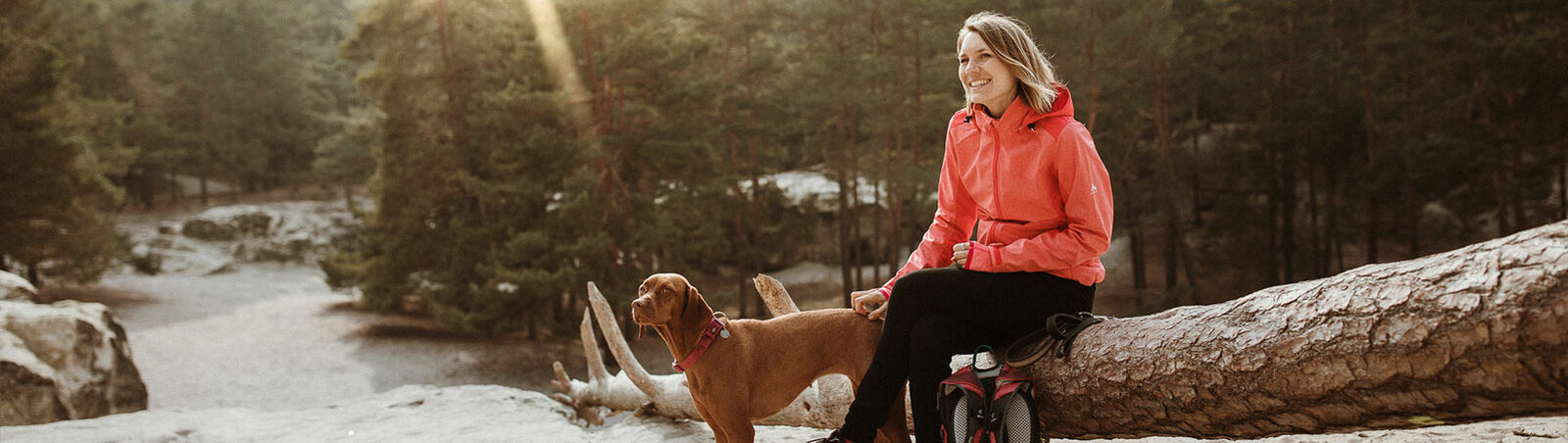 Hanna Busch in Wanderkleidung sitzt neben ihrem Hund in den Bergen auf einem Baumstamm.