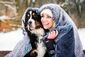 Ein Hund und eine Frau mit Mütze sitzen gemeinsam unter einer Wolldecke im Schnee.