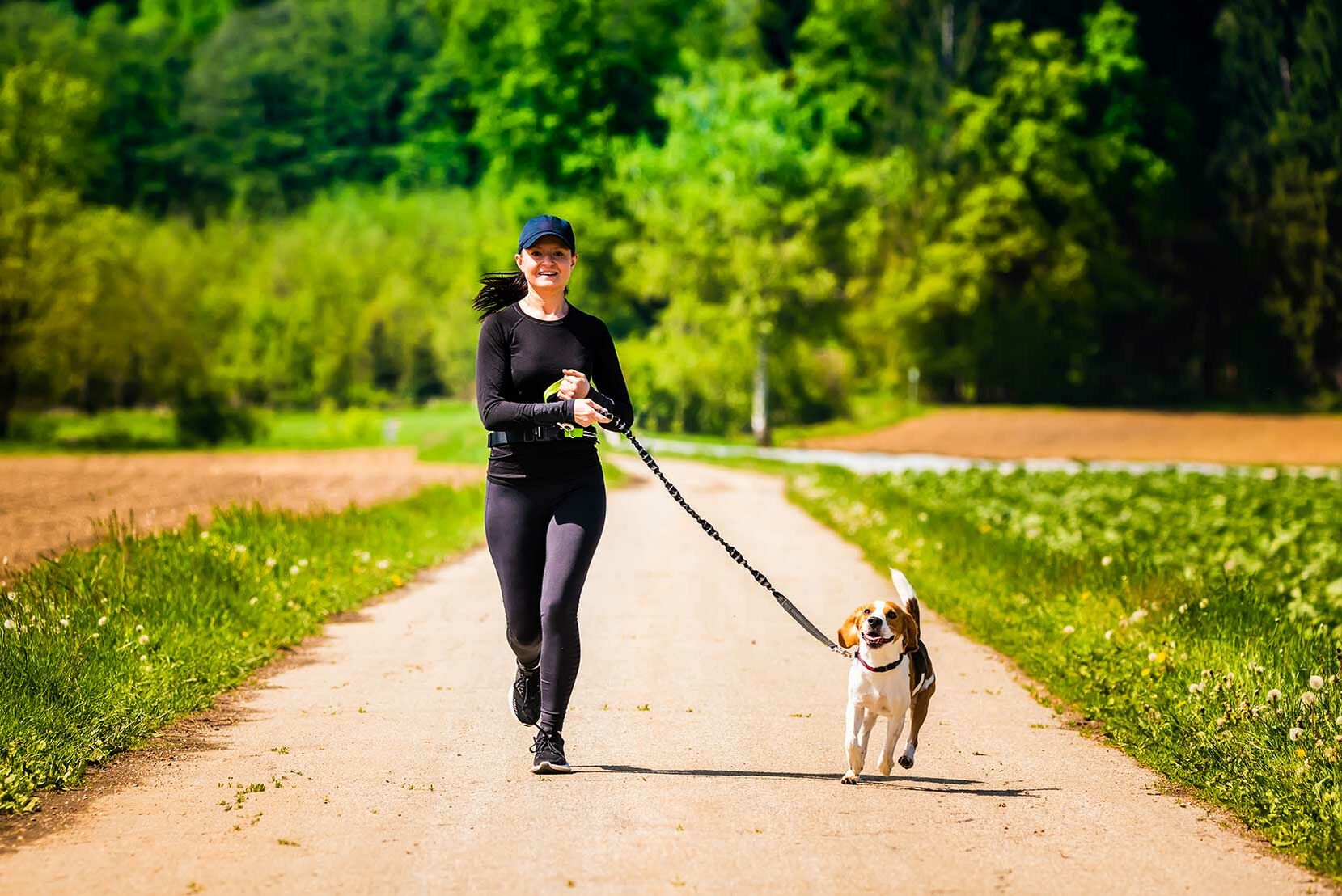 Frau joggt mit angeleintem Hund auf einem ländlichen Feldweg.