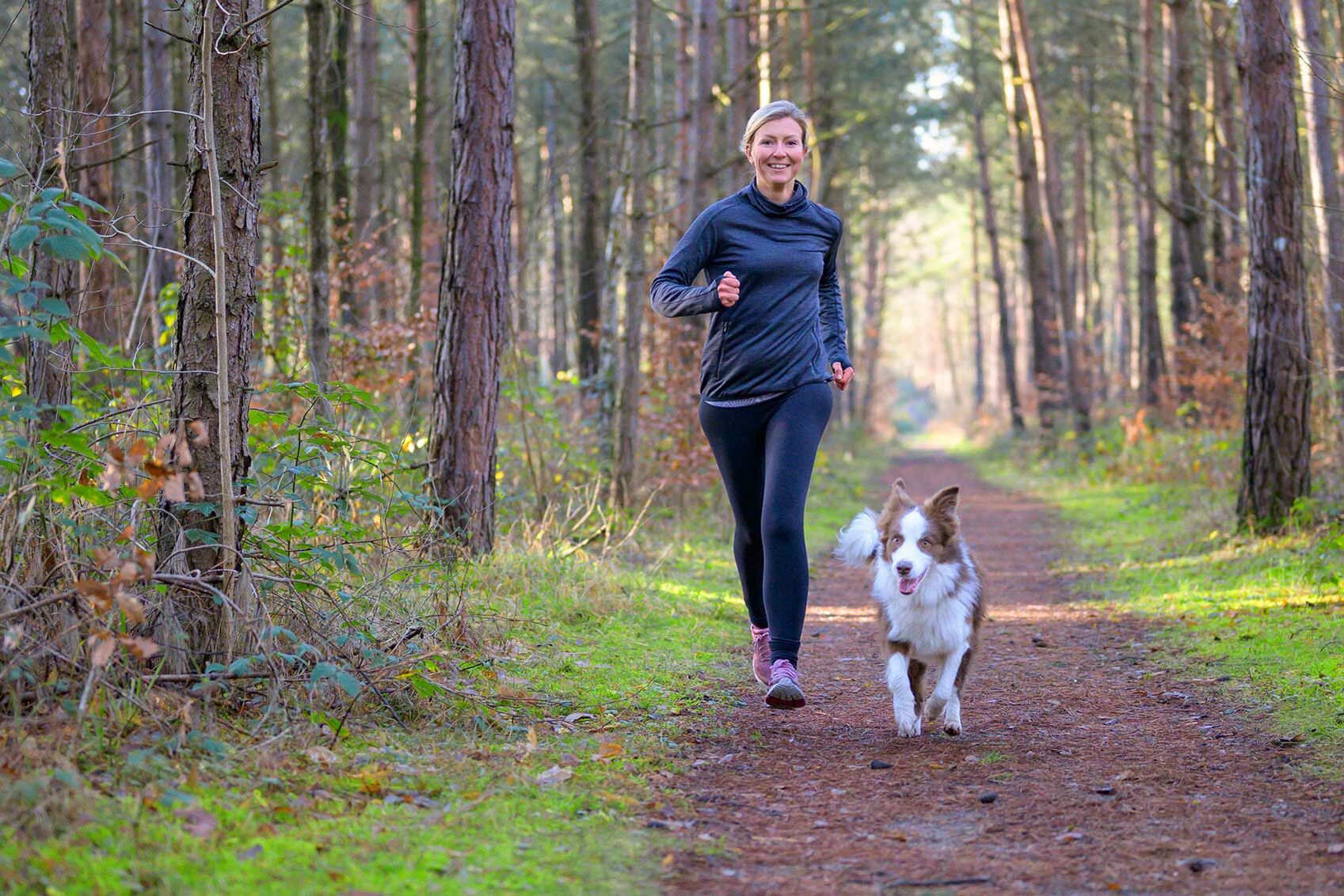 Eine Frau in Trainingskleidung joggt mit ihrem Hund auf einem Waldweg. 