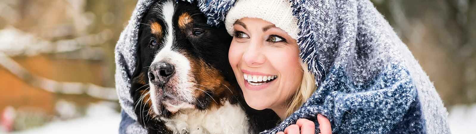 Ein Hund und eine Frau mit Mütze sitzen gemeinsam unter einer Wolldecke im Schnee.