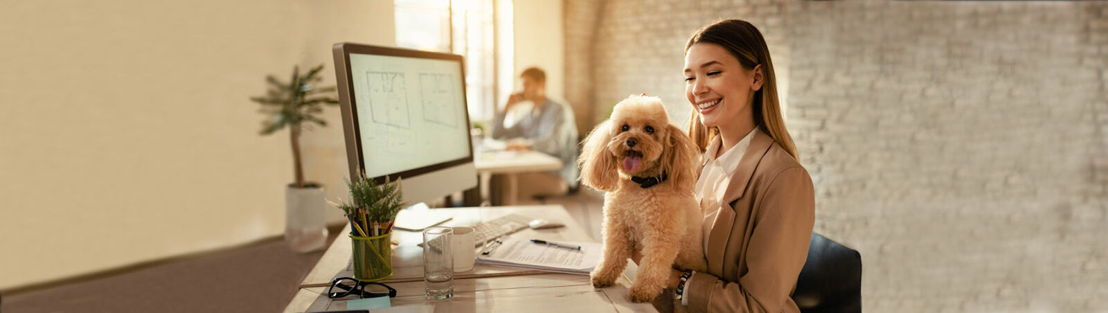 Eine Frau sitzt mit ihrem Hund auf dem Schoß an einem Schreibtisch im Büro.