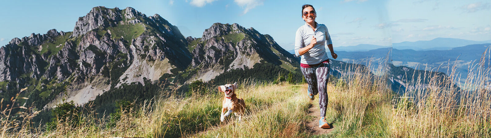 Eine lachende Frau joggt mit ihrem Beagle in einer Berglandschaft. 