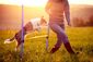 Ein Hund springt bei Sonnenaufgang über ein Agility-Hindernis auf einer Wiese und wird dabei von seiner Halterin angeleitet.