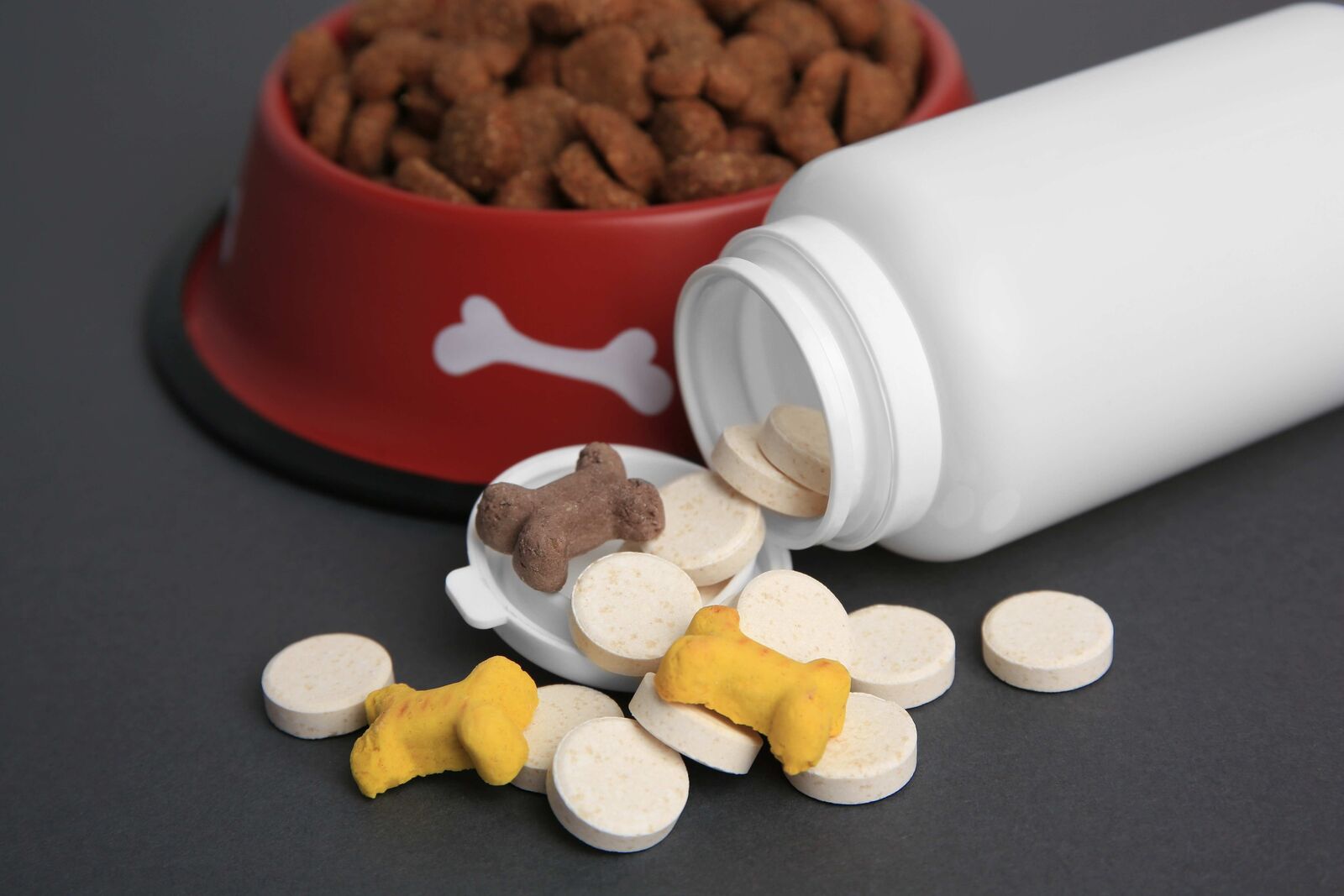 Im Hintergrund ein gefüllter Futternapf und im Vordergrund ein liegendes Gefäß, aus dem Tabletten fallen.